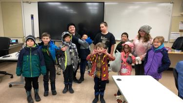 Elementary school children standing in a classroom with winter jackets, boots and toques on, making faces, smiling, watching.