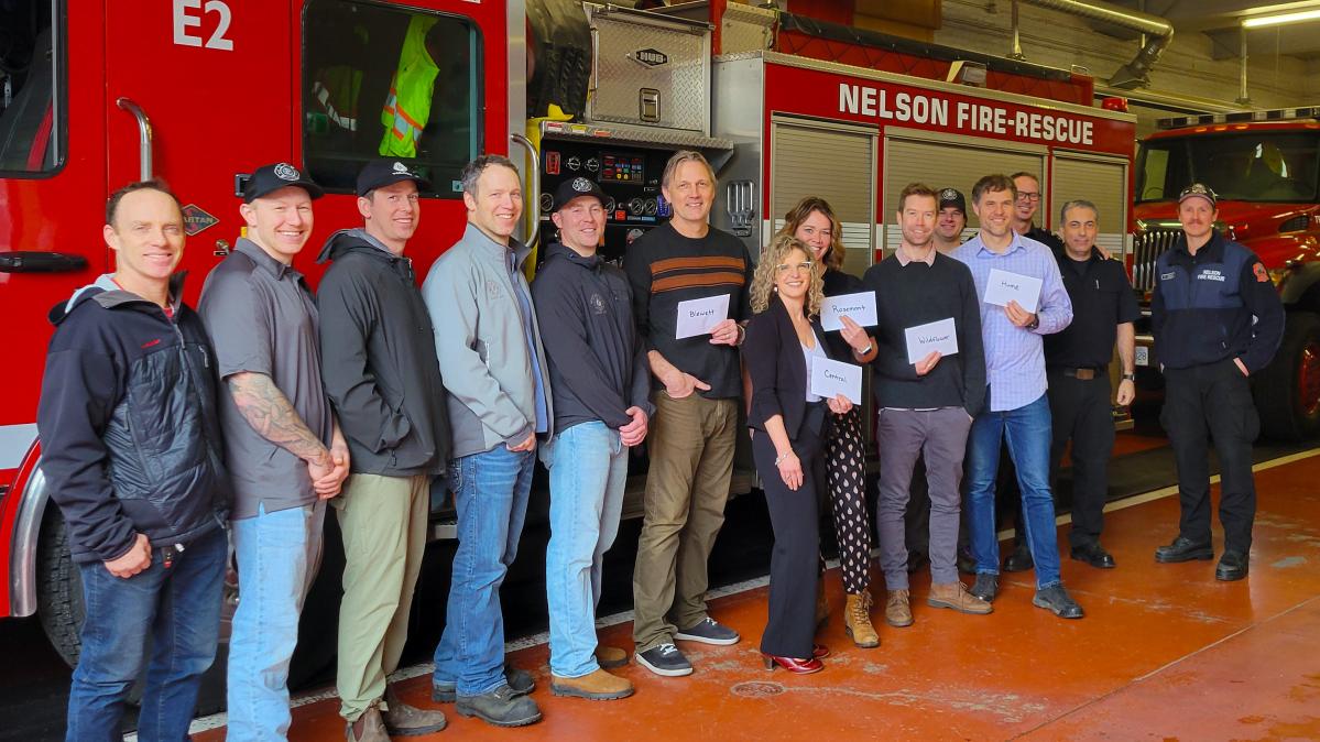 The Nelson Professional Firefighters Association presented $400 to each of five Nelson area schools in SD8 for assistance with food and meals for students and families. (front l to r) Nelson Professional Firefighters Scott Jeffry, Lucas Fortier, Adam Banilevic, Marc Thibault, Chris Thast; Blewett Elementary Principal Jordon Konken, South Nelson Elementary Principal Dawn Snell, Rosemont Elementary Principal Lindsay Mackay, Wildflower School Principal Nick Graves, Hume Elementary Principal Sacha Kalabis; and (rear l to r) Nelson Professional Firefighters Martin Ouellette, Greg Proctor, Rick Maida, Chas Misener A group of people lined up in front of a fire truck inside the bay of a fire department smiling, with some holding envelopes and some in fire fighter uniforms.