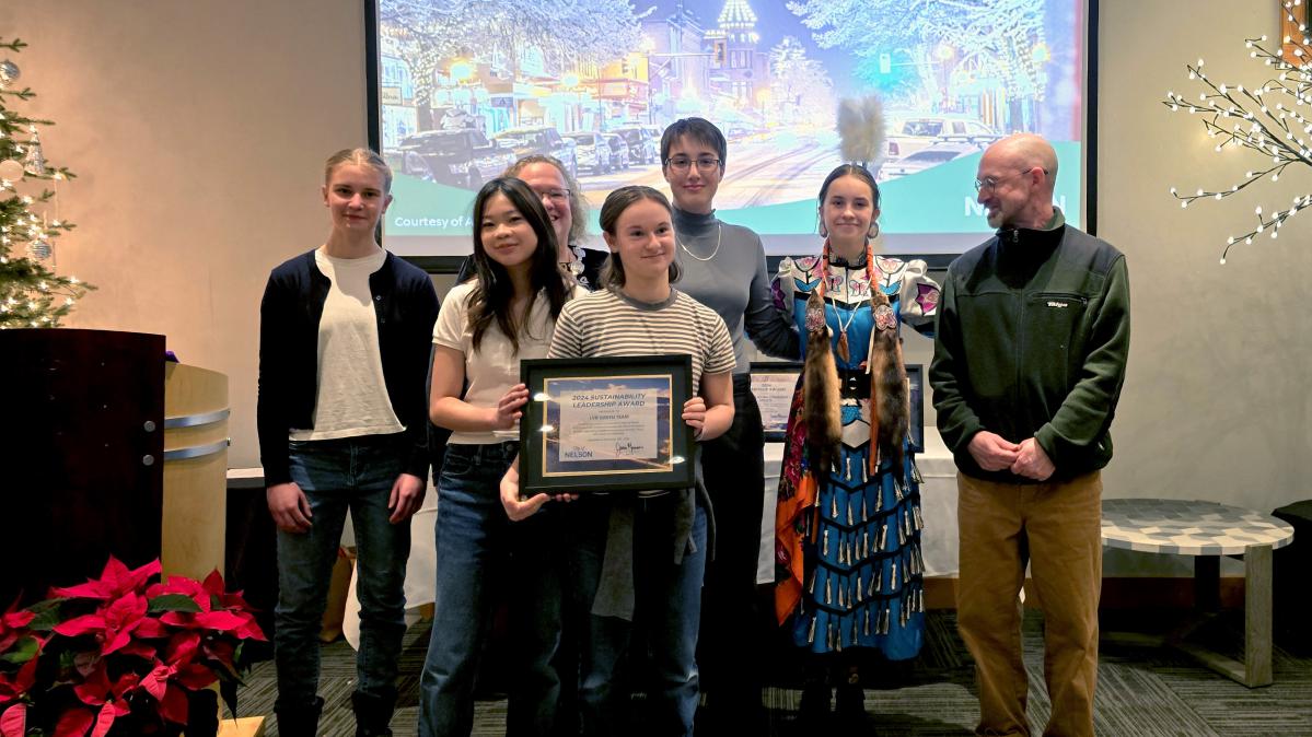 A group of young people holding a framed award posing in front of a screen and flanked to the right by an adult looking on.