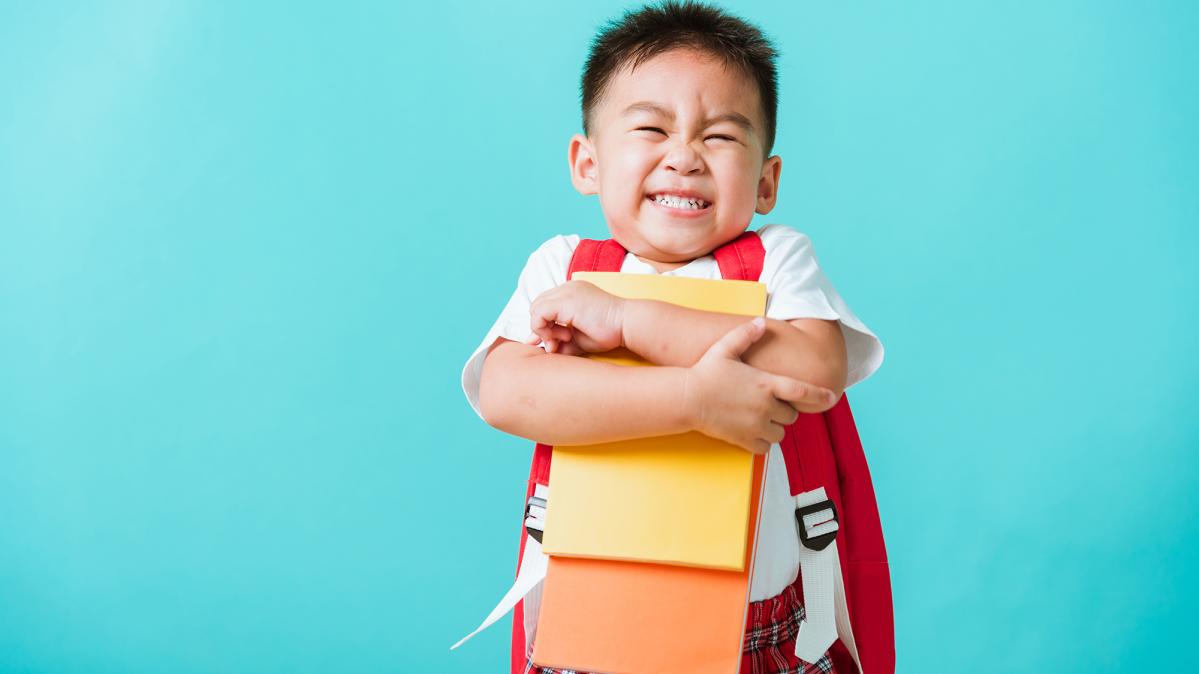 A preschool child with short black, spiky hair wearing a backpack and hugging some paper school supplies while he smiles in excitement.