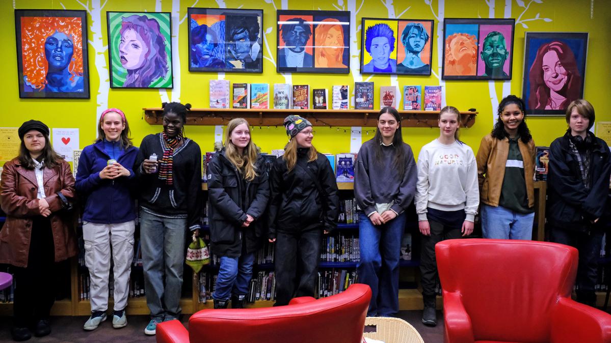 High school students stand in front of a yellow wall displaying their colourful artwork, with two red, leather chairs in the foreground. 