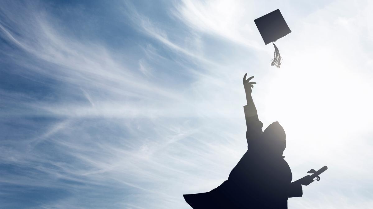 Grad 2025 Silhouette of a person in a graduation gown holding a diploma and throwing their tassled hat in the air against a blue, cloud painted sky.