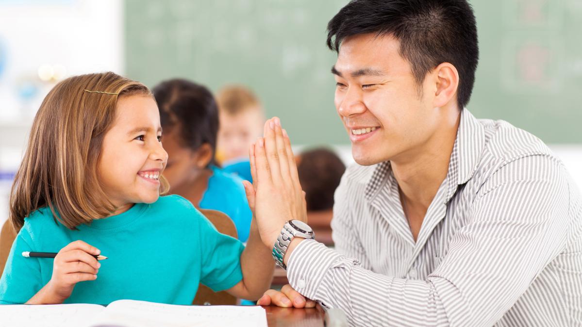 A young elementary student sitting at her desk high fives her male teacher who is crouched beside her. Both are smiling widely.