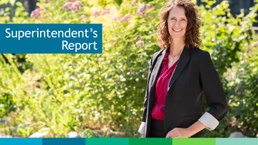 Smiling caucasian woman with brown curly mid-length hair in black blazer and magenta blouse standing in a relaxed pose in front of bright green and flowering bush in the sunshine.