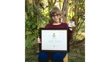 A young woman with wavy blonde hair wearing a burgundy sweater seated on a chair in a forest holding a medal and a framed certificate.