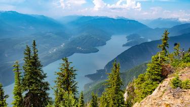 A large and long lake, mountains and blue sky and clouds seen from a mountain vista with evergreen trees in the foreground.