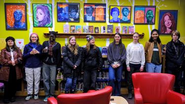 High school students stand in front of a yellow wall displaying their colourful artwork, with two red, leather chairs in the foreground. 