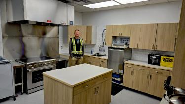 A carpenter wearing a reflective shirt stands in a newly renovated school kitchen.