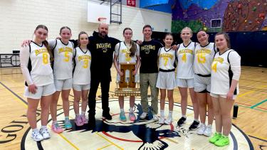 A highschool girls' basketball team stands with their coaches, holding a large trophy in the centre of a gymnasium.