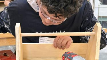 A teenager leans over a workbench working on a woodworking project with a red drill in the foreground and another teenager working on a project behind him.