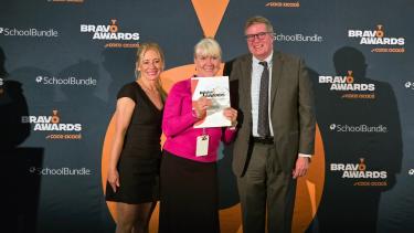 Two women in dresses and a man in a suit pose in front of a grey wall with the middle woman holding up a certificate.