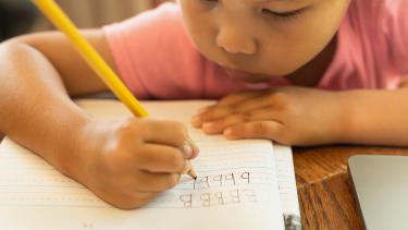 A closeup of a child holding a pencil and concentrating on writing letters of the alphabet in a lined notebook.