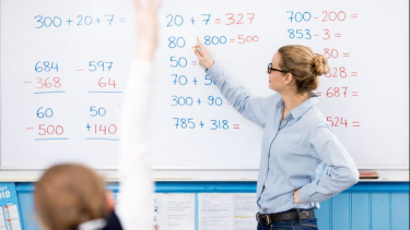 A female teacher shows math problems on a whiteboard while I young student in the foreground raises hand.