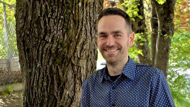 A man with short blonde-brown hair in a blue short-sleeved button-up shirt stands smiling in front of a tree in the sunshine.