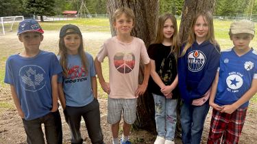 Six children stand outside in front of two trees with a school playground behind them.