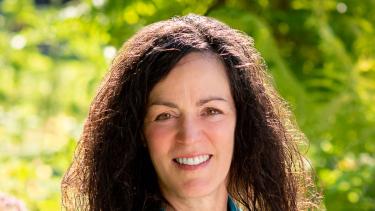 A woman with long dark hair wearing a short sleeved turquoise blouse smiles in front of green bushes outdoors in the sunshine