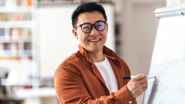 Man with black hair and black glasses teacher smiling writing on whiteboard in classroom