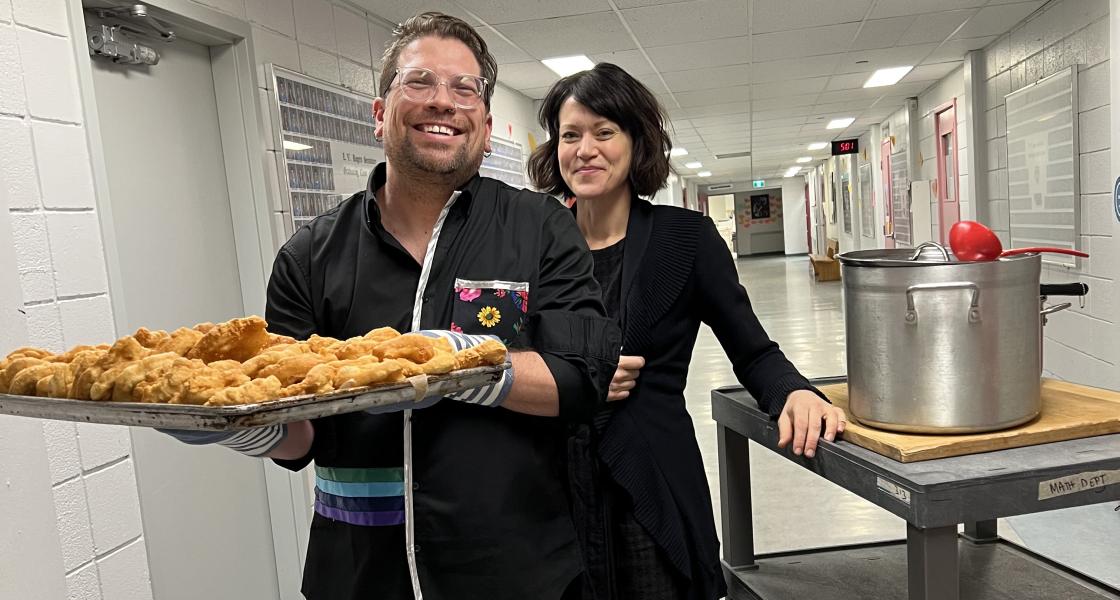 Smiling man in black chef's coat holds tray of baked goods in front of woman in black chef's coat who stands beside large silver stock pot.