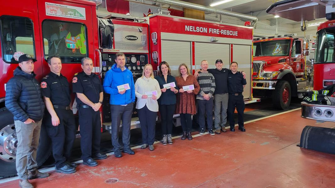 Row of firefighters in casual clothes and dress uniform along with civilians standing in front of a fire engine in a truck bay with four civilians holding cheques.