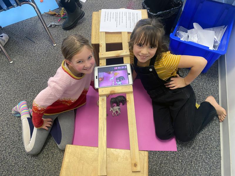 Two smiling girls sitting on grey carpeted floor on either side of a computer tablet with purple video image showing.