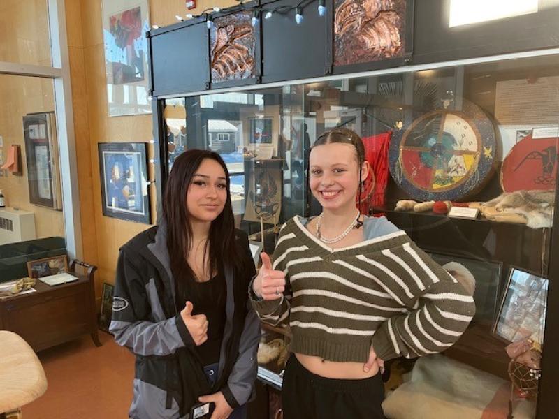 Two young women giving thumbs up while standing in front of an Indigenous art display in their high school foyer