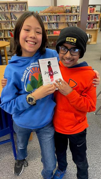 Two smiling children with their arms around one another, holding up a book in a library