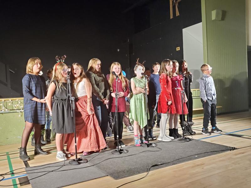 Children poised to sing, standing in front of a grey mat in a school gymnasium.