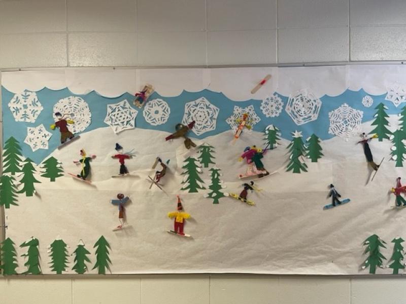 Large rectangular poster showing paper cutouts of skiers on a treed slope with paper snowflakes on a blue sky above the paper skiers with the entire poster mounted on a white, brick wall., 