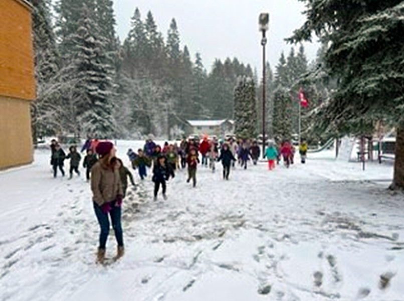 A young, female adult wearing a burgundy toque leading school children in play in a snowy winter schoolyard