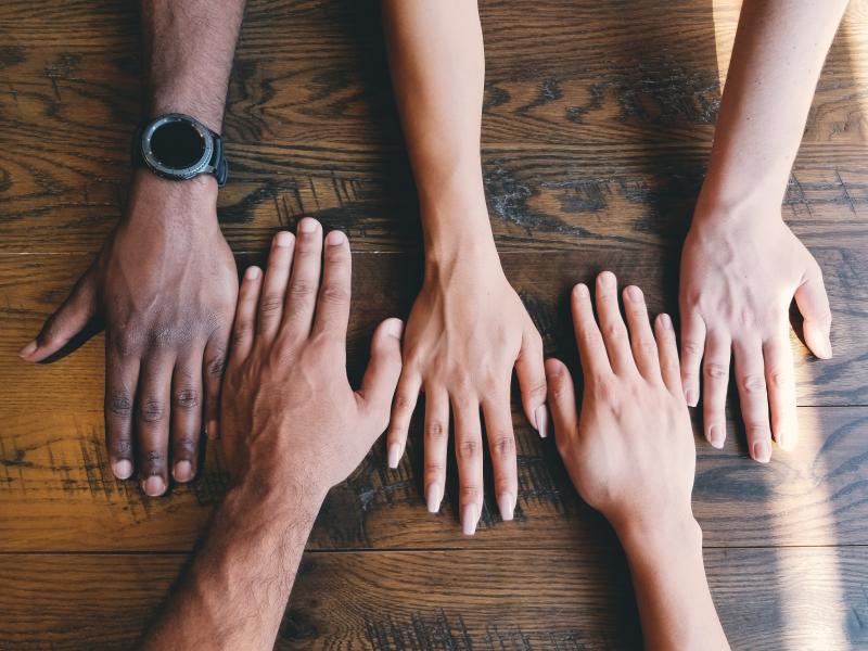 Multi-racial and multi-gendered hands palm-down on a wooden table with fingers alternating facing top and bottom of the image. One hand shows a watch on the wrist.