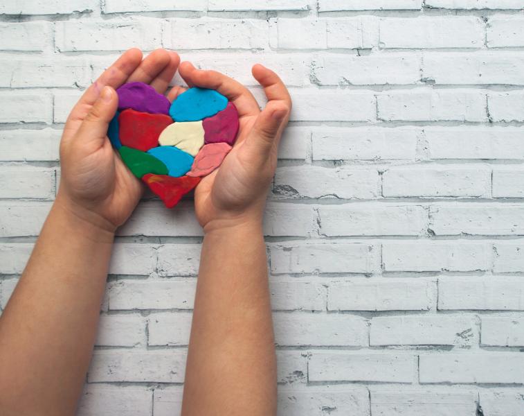 Child's hands cupping a colourful plasticine heart against a white brick wall.