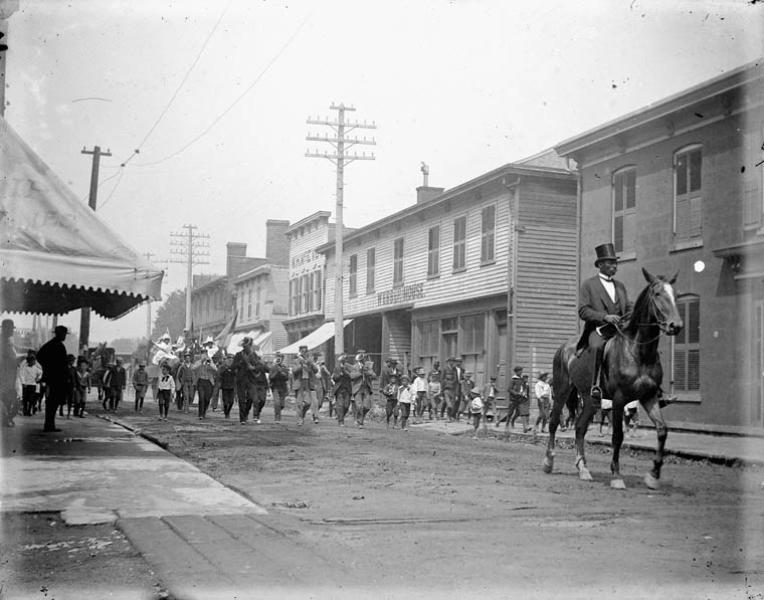 A black and white historic photo of a man in a top hat riding a horse at the front of a parade of people