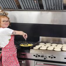 A woman with red hair in a scrunchie on top of her head and wearing a red, flowered apron stirs scrambled eggs in a pan beside an array of pancakes on a commercial grill.