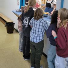 Children line up at a school kitchen serving window while adults serve them breakfast.