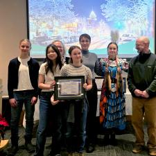 A group of young people holding a framed award posing in front of a screen and flanked to the right by an adult looking on.