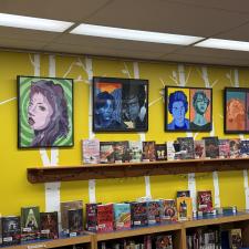 Colourful Warholesque portraits hanging on a yellow wall above a bookshelf in a library.