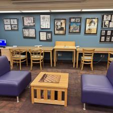 Two mauve armless couches facing a checkerboard table with wooden chairs and a bench as well as artwork on the walls in the background.