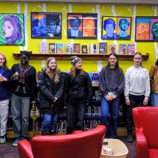 High school students stand in front of a yellow wall displaying their colourful artwork, with two red, leather chairs in the foreground. 