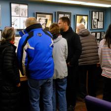 A group of people in winter jackets stand facing a wall with various artwork hanging on it.
