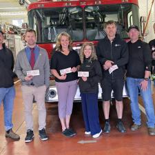 A group of people stand in front of a fire truck inside a fire department, holding cheques in front of themselves.