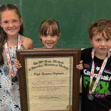 Three children stand in front of a table and chalkboard holding a framed award from 1924
