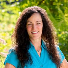 A woman with long dark hair wearing a short sleeved turquoise blouse smiles in front of green bushes outdoors in the sunshine